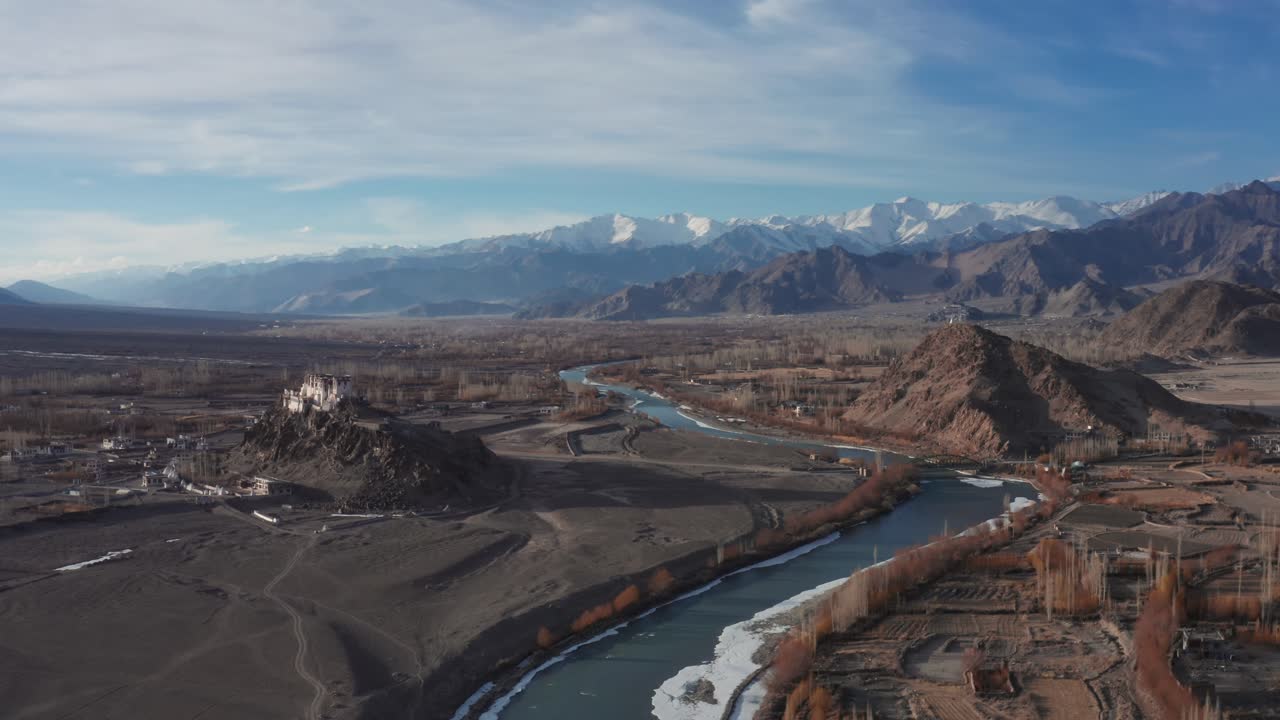 Luftfoto fra øst af Stakna-klosteret på en klippe med Indus-floden, der snor sig gennem en gold dal, landsbyer og fjerne snedækkede Ladakh-bjerge