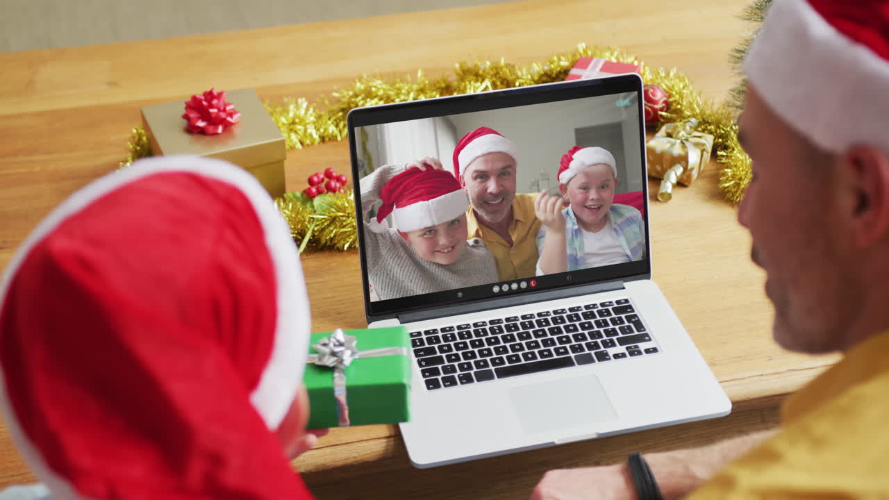 padre y hijo caucásicos con sombreros de santa usando una computadora portátil para una videollamada de navidad con la familia en la pantalla