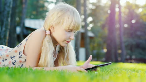 blonde girl 6 years playing on the tablet emotionally reacts lying on the lawn in the backyard of th