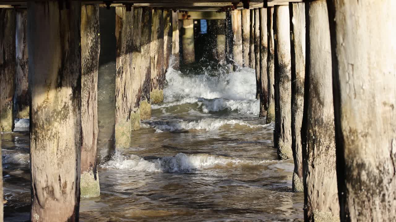 Dynamic waves crash against a weathered wooden pier in Bellarine, Victoria. Sunlight casts dramatic shadows, creating a rhythmic, natural scene