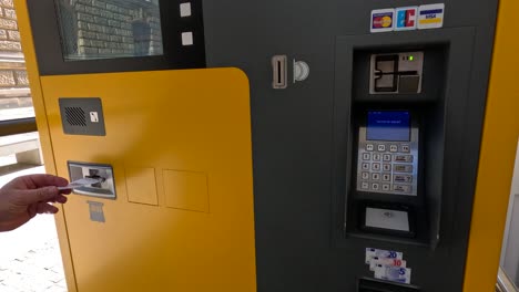 person inserts card into yellow outdoor ticket machine payment terminal in daylight, close-up