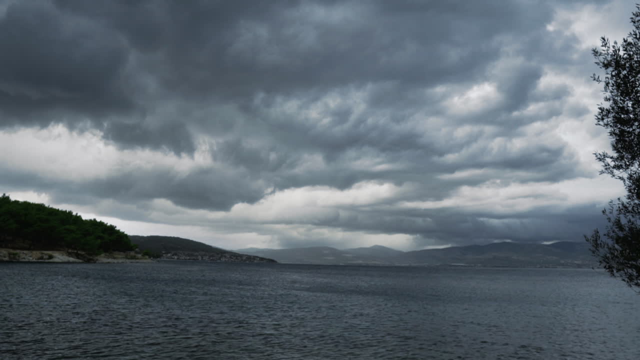 Timelapse of dramatic storm clouds gathering over coastal landscape. Squalls, gusts of wind visible on water. Dense greenery on rugged shoreline. Wide angle. Croatia.