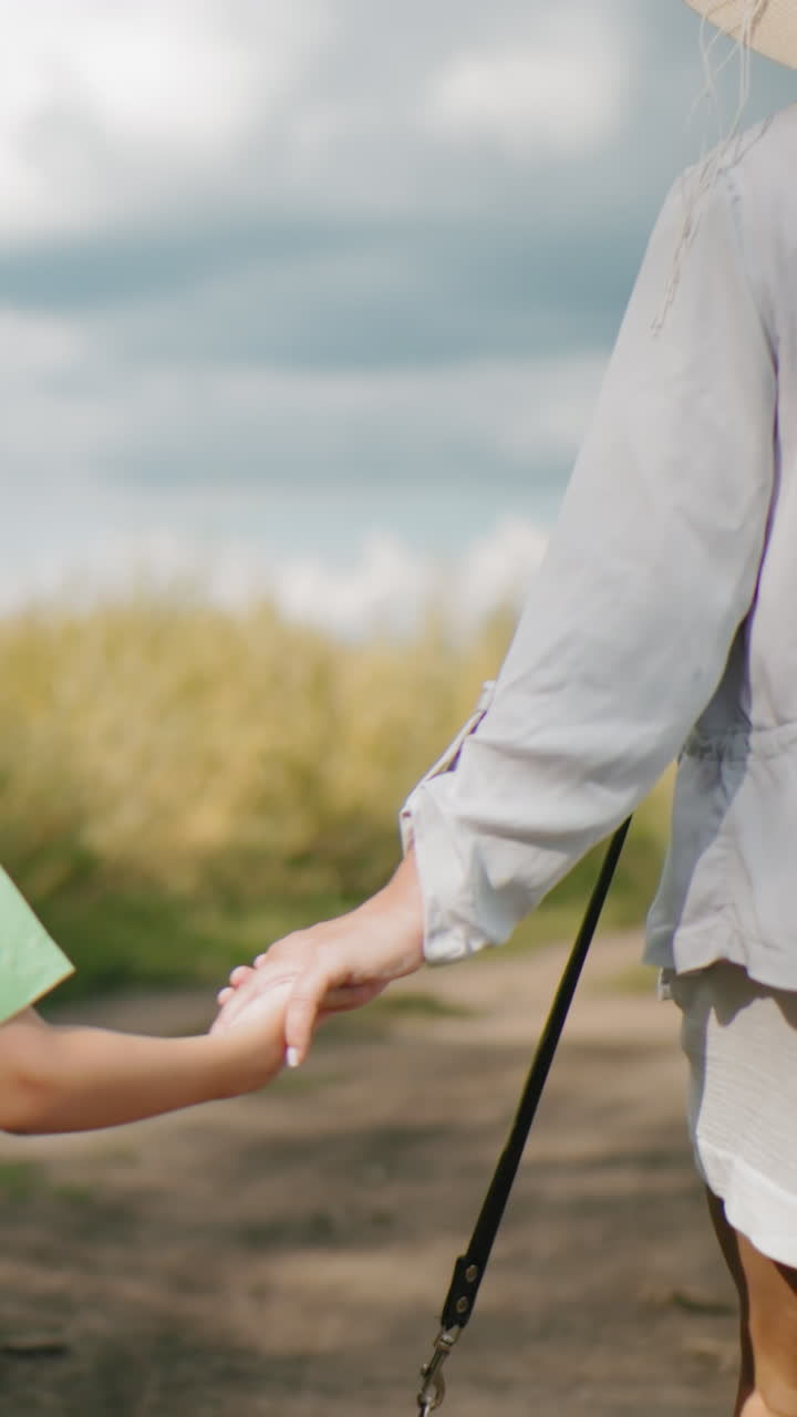 모자를 입은 여자가 아들의 손을 들고 분홍색을 입은 소녀가 길을 따라 걸어가고, 부분적으로 구름이 가득한 하늘 아래 초록색으로 둘러싸여 있으며, 그들 뒤에 바구니를 들고 있는 다른 사람의 부분적인 시야가 있습니다.