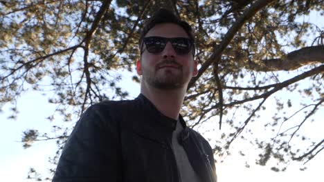 young bearded man with sunglasses and leather jacket stands in sunshine under a tree