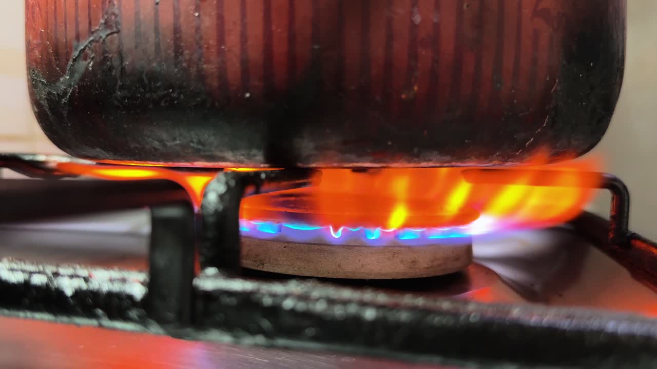 Close-up de um queimador de gás de cozinha acendendo com chama azul enquanto uma mão ajusta o fogão ao lado de uma panela aquecida, em luz interna quente e pouca profundidade de campo