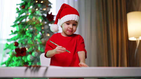child with santa hat crafts. a child in a santa hat sits at a table with a green pencil, focused on holiday crafts by a decorated tree