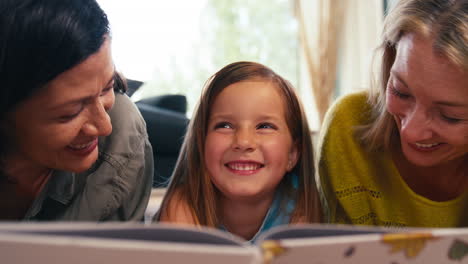 primer plano de una familia del mismo sexo con dos madres e hija acostadas en el suelo leyendo un libro en casa juntas