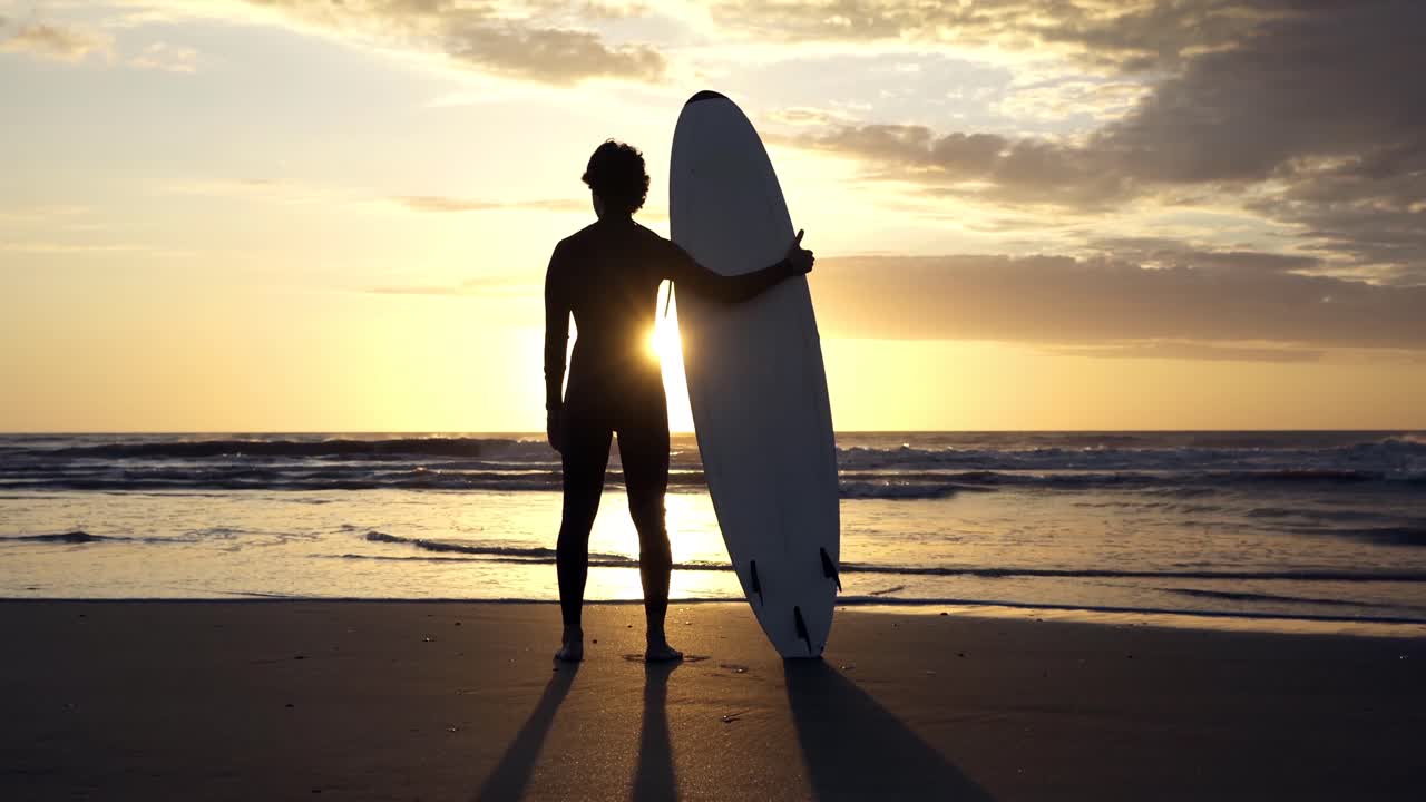 Silhouette di surfista in piedi sulla spiaggia con tavola da surf all'alba
