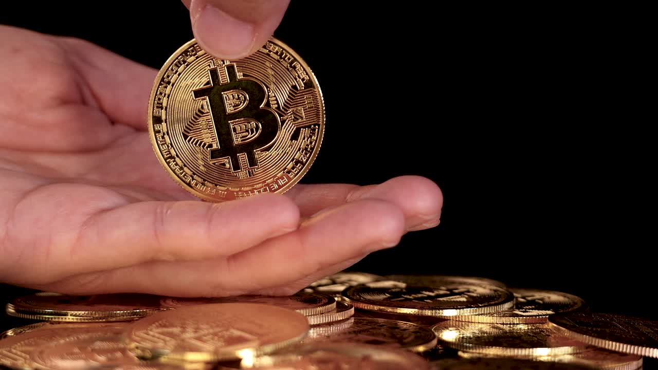 Human hand offers golden bitcoin coin above pile, dramatic lighting, close-up, black background