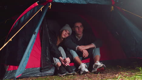 romantic moment as couple sits close together inside tent during night camping in forest, sharing quiet time with heads leaned and hands gently touching, surrounded by soft light