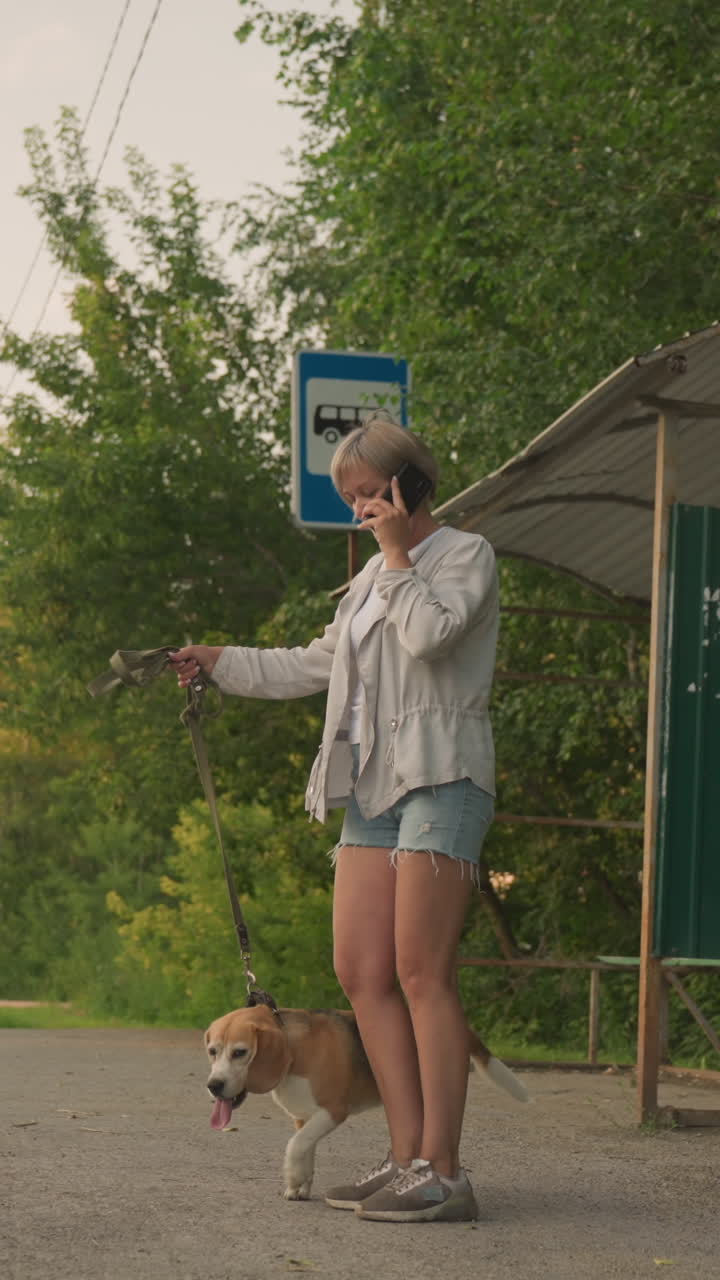 el dueño del perro de pie en la parada de autobús en una zona rural sosteniendo al perro en la correa mientras habla por teléfono mirando contemplativo, los alrededores incluyen árboles verdes, parada de autobuses, refugio y carretera con coches que pasan