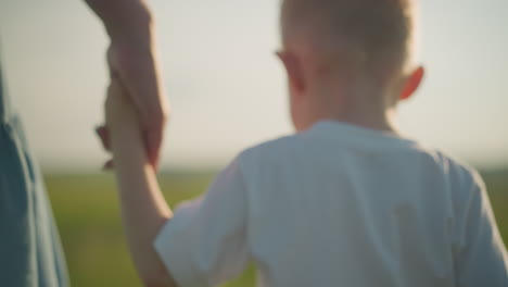 un close-up de un niño pequeño en una camisa blanca de la mano con una mujer en un vestido azul, su cara no es visible, mientras caminan a través de un campo de hierba iluminado por el sol, borroso
