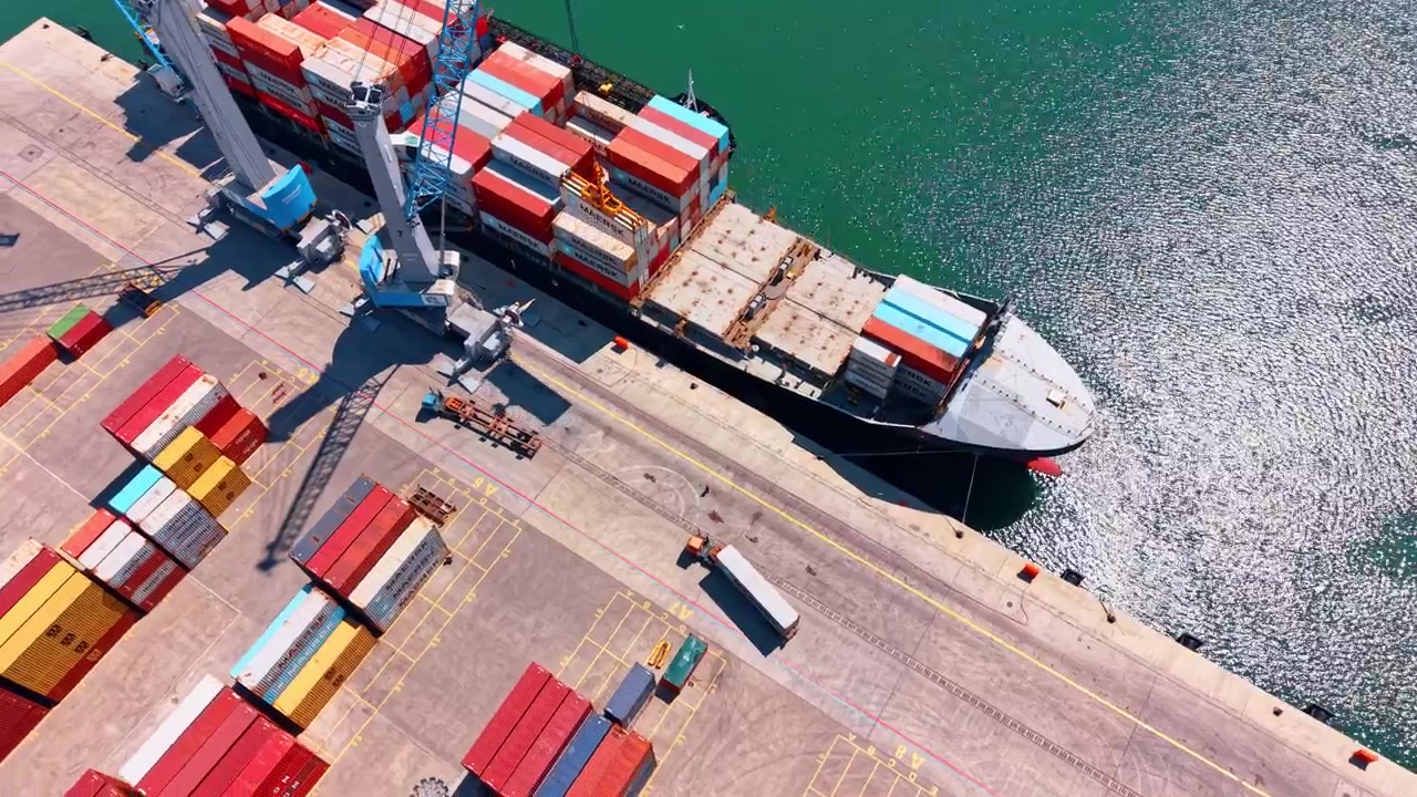 Burgas, Bulgaria, 1 May 2026: Cargo ship loading containers beside mobile harbor cranes. Oblique aerial view of container ship at quay with two cranes, stacked containers and sunlit harbor water.