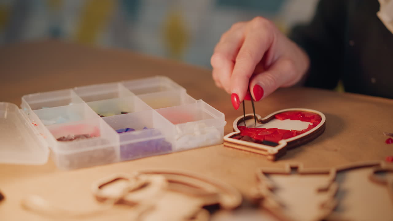 Closeup hands crafting christmas heart present, artisan picking tiny beads with tweezers on wooden table, red nails, warm lamp glow, DIY gift tag scene, detailed texture and soft bokeh
