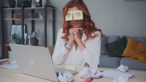 caucasian woman sleeps sits on desk with laptop with paper stickers on eyes