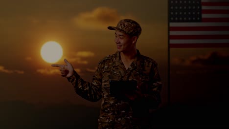 asian man soldier using a tablet and pointing to side while standing with flag of the united states, sunset time