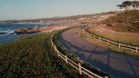 Group-of-cyclists-riding-on-a-curved-road-at-the-seaside-with-scenic-view-of-the-sea,-aerial-tracking-shot