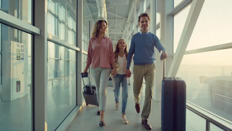 cheerful parents and their teen cute daughter walking the passage at the airport with suitcases happily