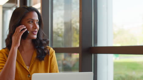 Mature-Businesswoman-Working-On-Laptop-At-Desk-In-Office-Taking-Call-On-Mobile-Phone