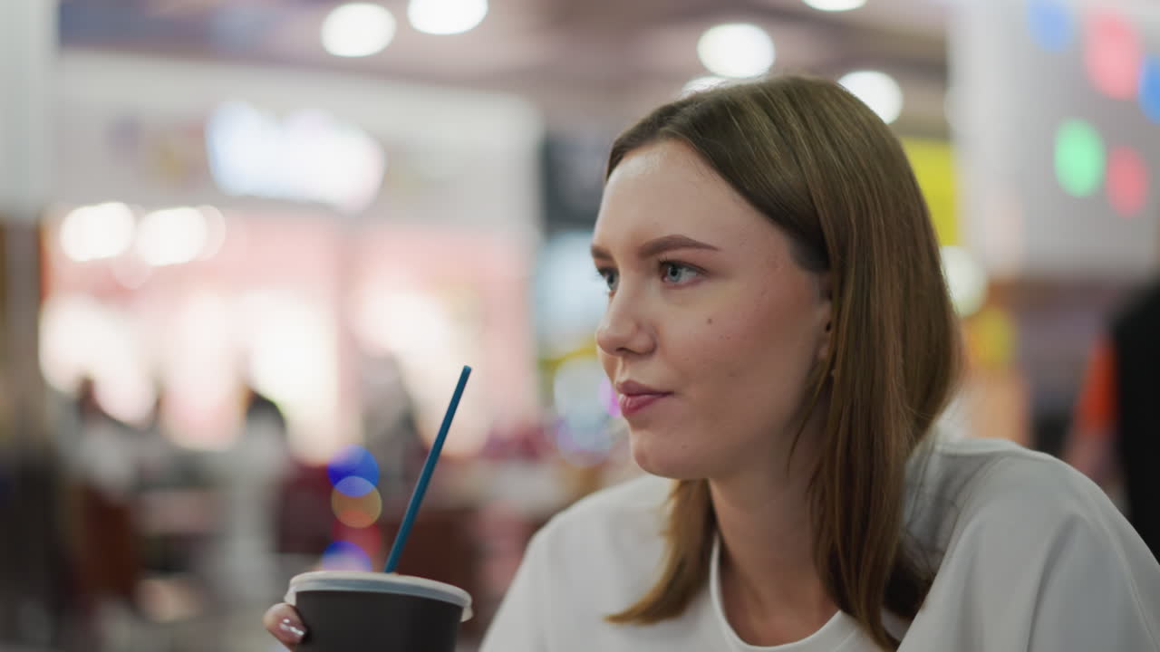 joven beba de una taza a través de la paja en la concurrida zona de comidas del centro comercial, su atención en la bebida en medio del colorido y borroso fondo del animado centro comercial