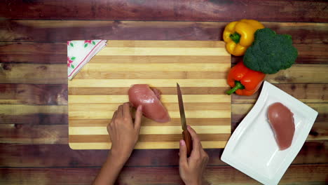 a top view of taking slice of a chicken from a try to cutting board for making slices, two big yellow and red capsicums, a boneless chicken piece, grater and a green broccoli on the table