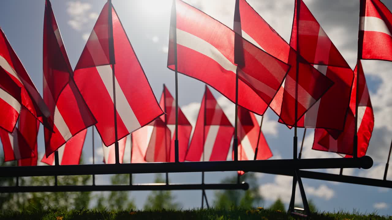 Wide establishing of Latvia flags blowing gently in wind with historic buildings behind, backlit sun light bloom
