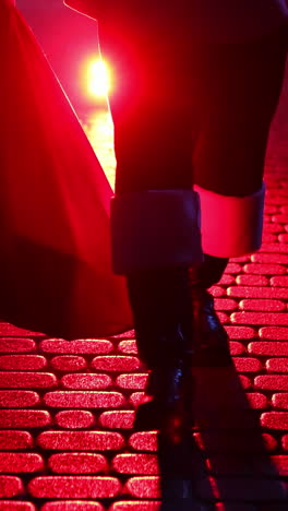 legs of santa claus. father frost carrying presents at christmas in red bag outdoors. black boots of santa walking on a pavement at dark on snow background. red light. vertical video