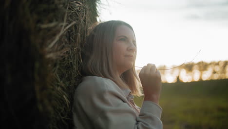 close-up of woman leaning against hay bale, holding a strand of straw while gazing into distance, sunlight creates a golden glow, highlighting her serene expression