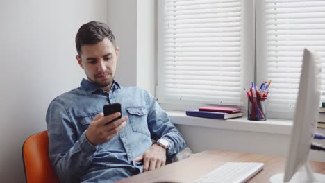 Joven-Oficinista-Usando-Su-Teléfono-En-La-Oficina-Sentado-En-La-Mesa-Con-Computadora,-Teléfono-Y-Taza.-Filmado-En-4k