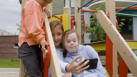 Camera-zooming-on-a-little-girl-with-down-syndrome-and-her-friends-making-a-selfie-with-smartphone-in-the-park-on-a-windy-day