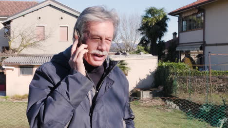old man with gray hair mustache with blue winter jacket talks and laughs during a phone call outside the houses with palm trees on the background in a sunny winter day - slow motion
