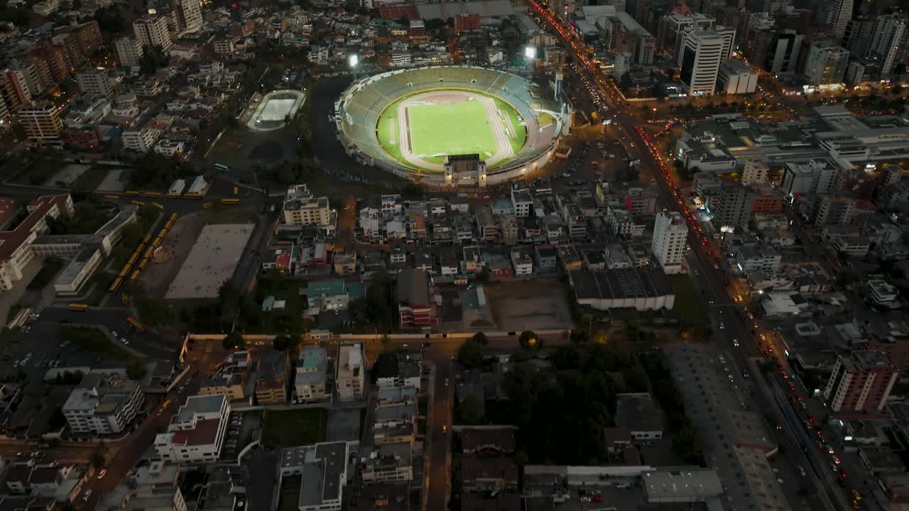 toma aérea inclinada que muestra el paisaje urbano de la ciudad de quito en ecuador con estadio de fútbol iluminado durante el juego y nubes místicas entre montañas en segundo plano