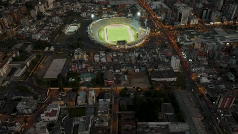 aerial tilt up shot showing cityscape of quito city in ecuador with lighting soccer stadium during game and mystic clouds between mountains in background