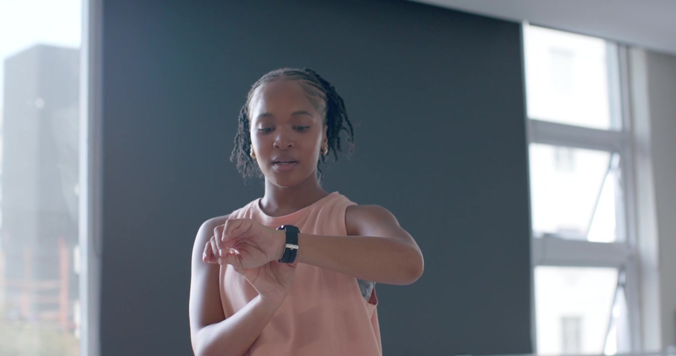 African woman ending shoulder raise in gym, placing hex dumbbell, checking smartwatch timing