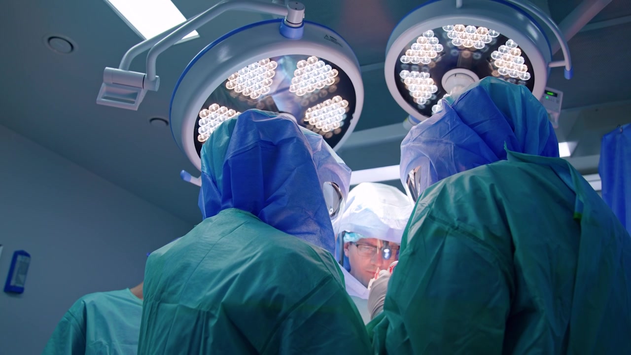 Vista posteriore di medici che usano un dispositivo elettrico che produce fumo. Medici con caschi protettivi operano sotto le lampade rotonde. Prospettiva dal basso