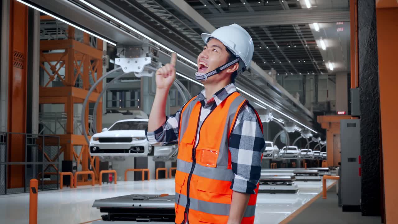 안전 헬을 입은 아시아 남성 엔지니어의 측면 뷰 생각하고 주위를 둘러보고 ev 자동차 공장에서 서있는 동안 지표손가락을 들어 올립니다.