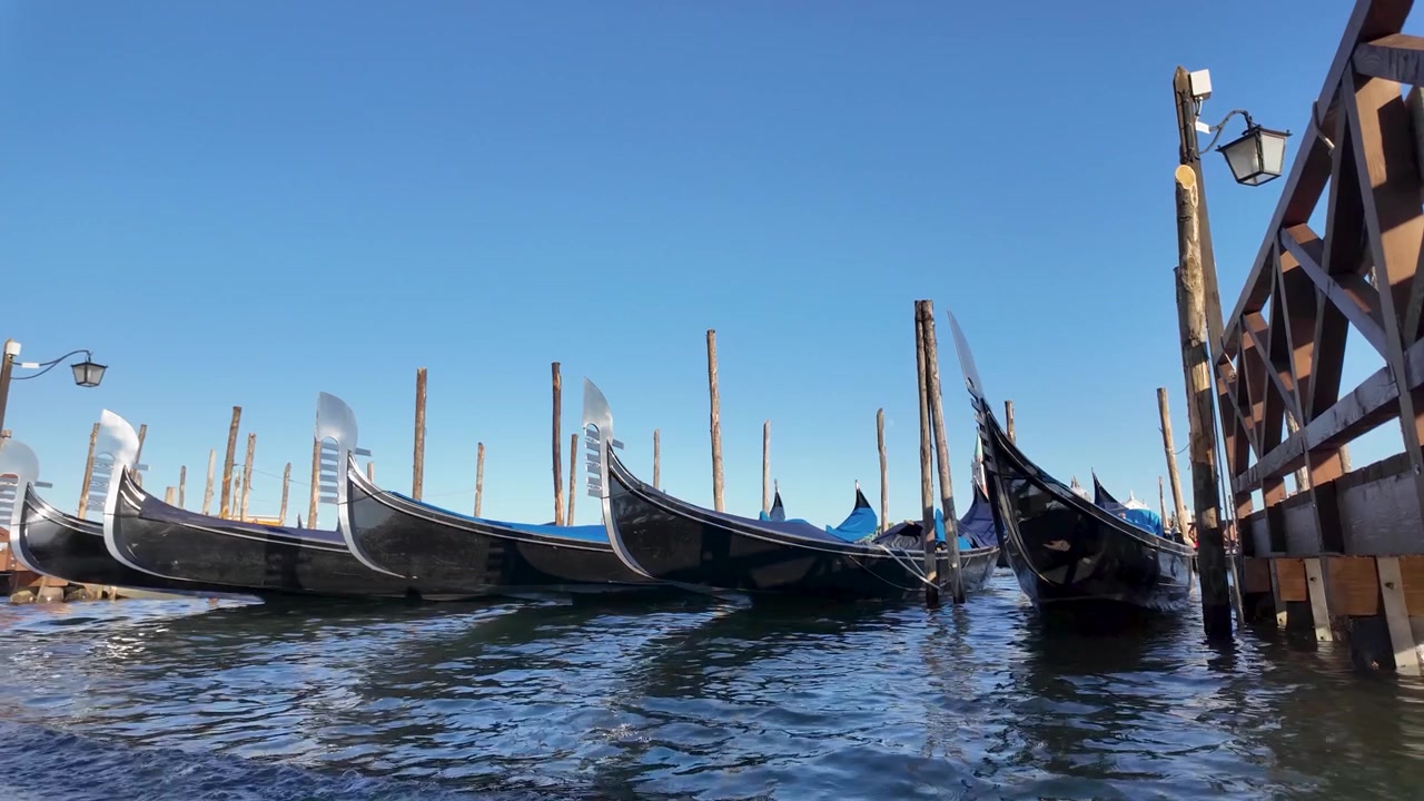Vue au ralenti de gondoles noires traditionnelles amarrées le long du front de mer à Venise, avec des poteaux d'amarrage en bois, l'eau ondulante du canal et un bâtiment vénitien historique sous un ciel bleu clair.
