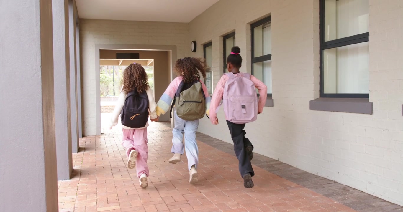 Três meninas afro-americanas caminhando na escola, a menina do meio liderando uma corrida em direção a um arco, mochilas
