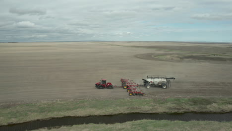 aerial scenic view of red industrial tractor seeding machine pulling fertilizer in flat farm field in expansive rural countryside on cloudy day, vanguard, saskatchewan, canada