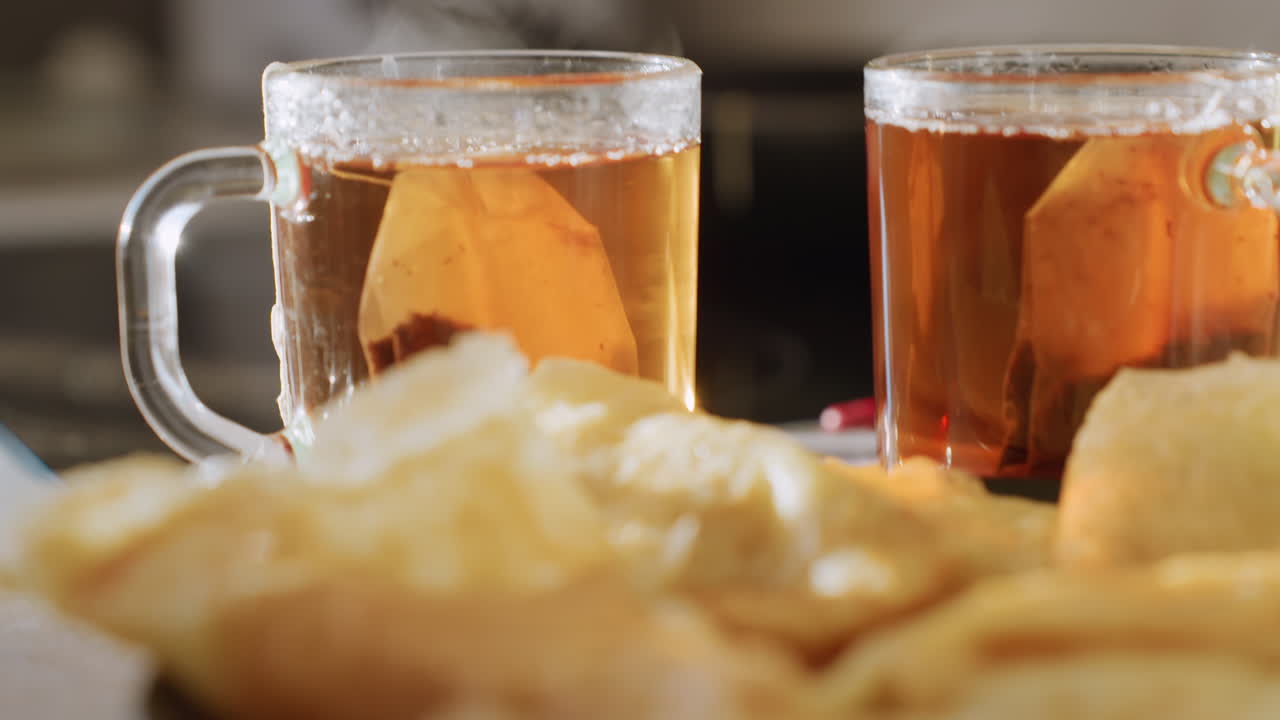 Close up of freshly cooked pancakes neatly arranged on plate with two cups of hot tea visible in background, golden texture of pancakes highlighted by warm light