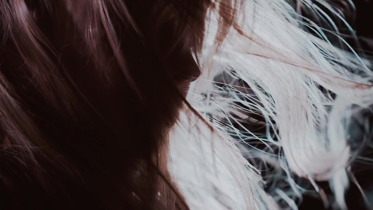 Primer plano melancólico de una cabeza girada con el cabello barrido hacia afuera por la brisa o el movimiento, mientras una fuerte luz lateral y de contorno ilumina hebras individuales, creando reflejos brillantes, contraste suave y