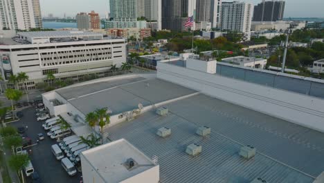 forwards fly above town development, tilt up and focus on american flag with modern downtown buildings in background. miami, usa