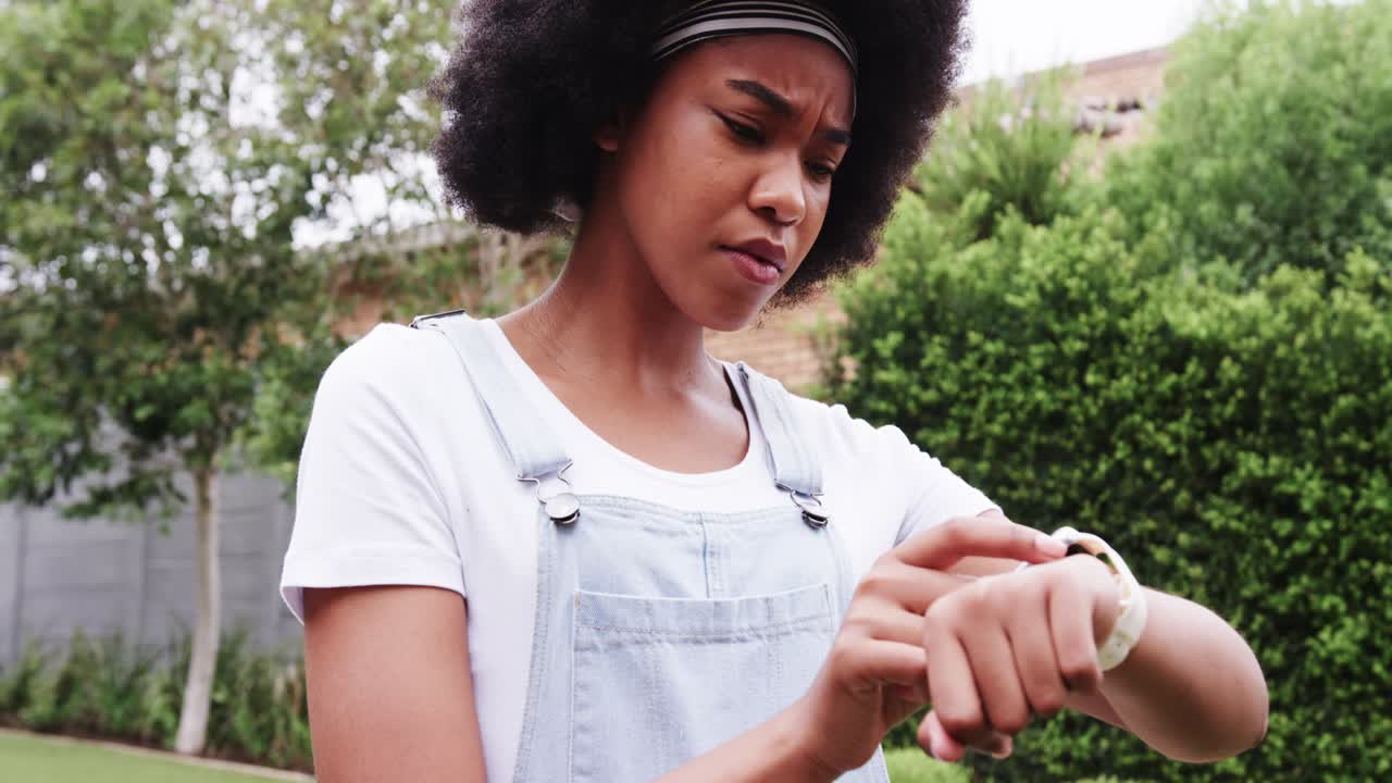 Adult woman inspecting and tapping white wrist device in backyard wearing denim overalls