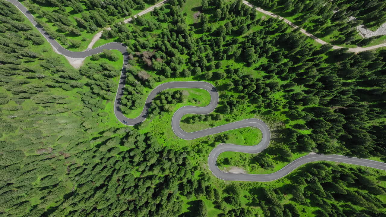Cinematic drone view of a car driving along the iconic snake road of Giau Pass in the Dolomites, framed by rolling green hills