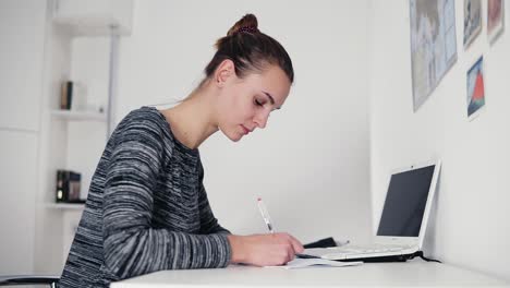 hermosa joven sentada junto a la mesa y escribiendo una carta usando su bolígrafo. abrir la computadora portátil en la mesa. mujer de negocios haciendo