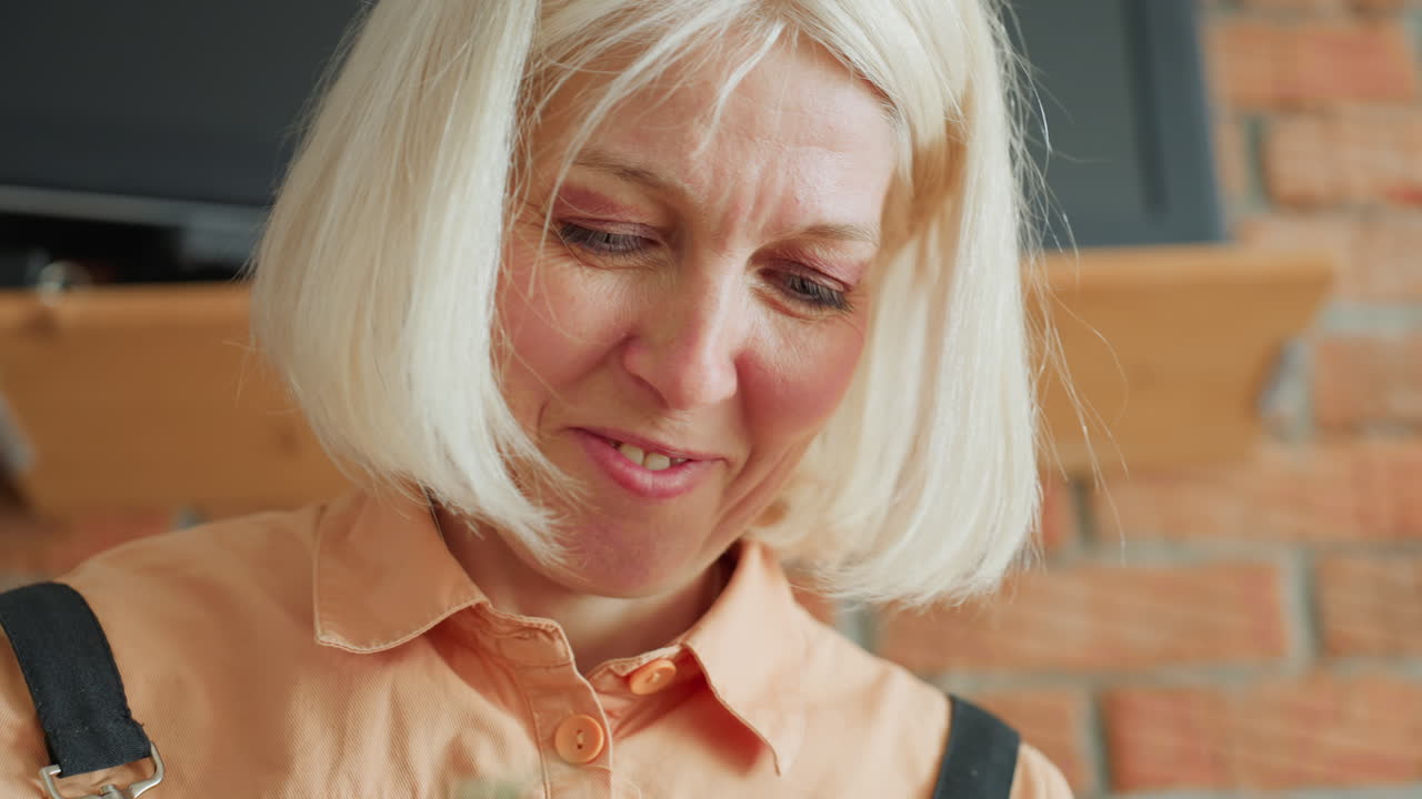 Decorator with blonde hair in orange shirt and black apron smiling gently while working on creative handmade project, close-up face portrait showing concentration and satisfaction in cozy indoor workspace