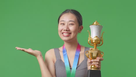 close up of asian woman with a gold medal looking at a gold trophy in her hands, smiling, and pointing to side on green screen background in the studio