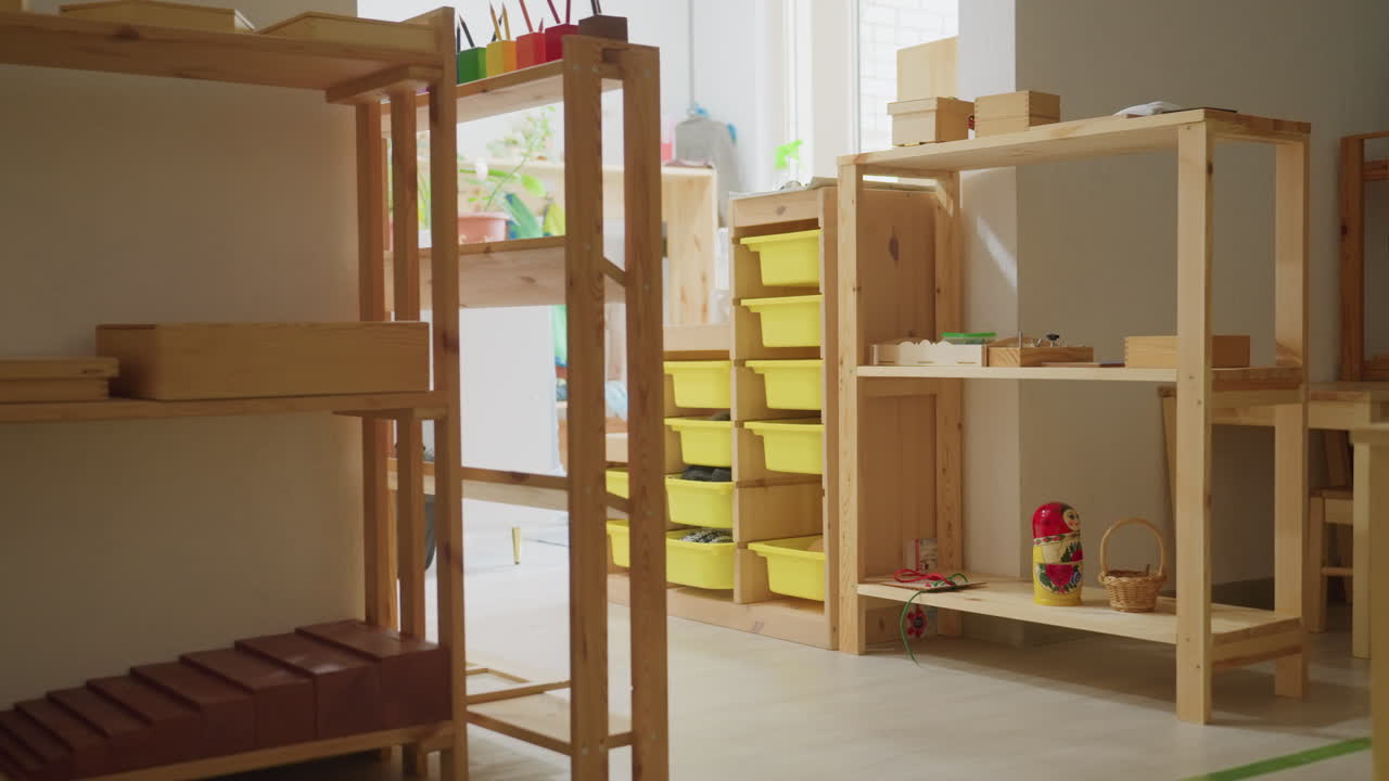Bright childcare classroom interior with wooden Montessori furniture, educational toys, colorful trays, and geometric blocks arranged neatly on shelves under daylight entering through large windows