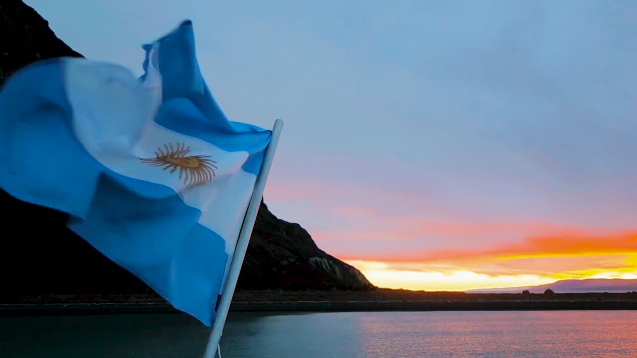 Argentinian flag blowing in wind at sunset by water and dark mountain silhouette