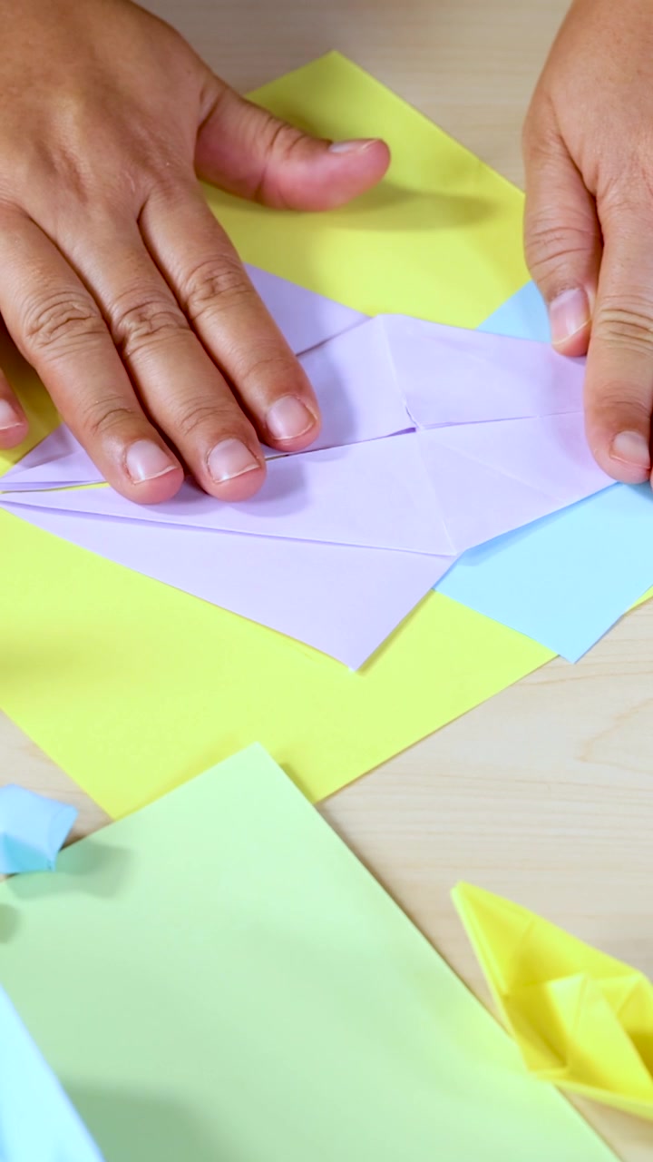 Una persona piega carta origami pastello su un tavolo di legno chiaro, illuminazione naturale, prospettiva ravvicinata.